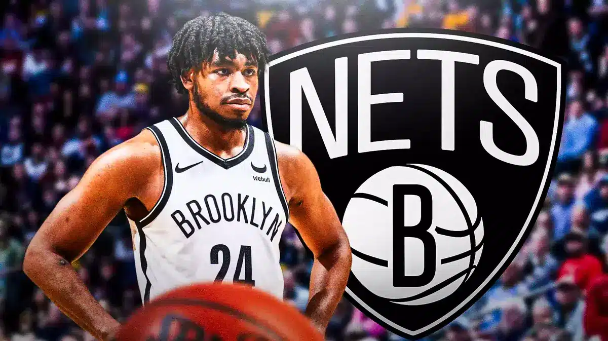 Cam Thomas in middle of image looking stern, Brooklyn Nets logo, basketball court in background
