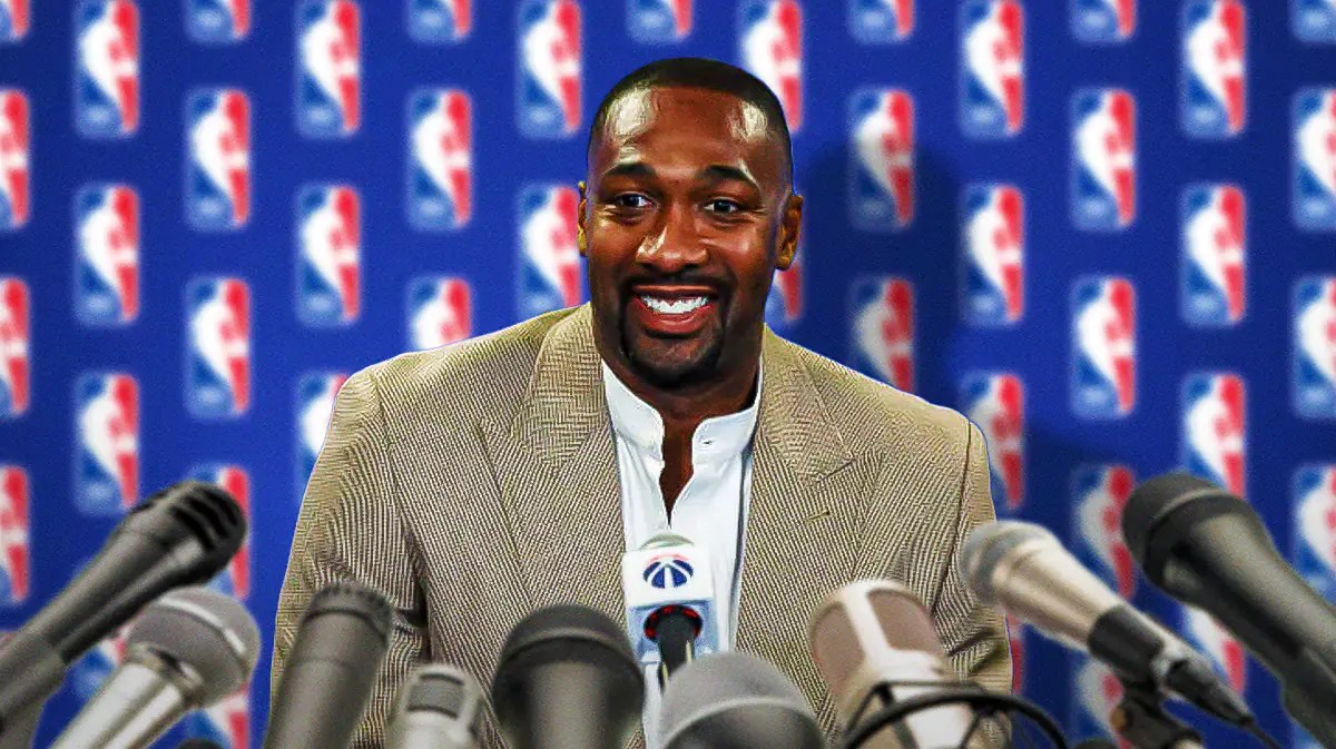 Former player Gilbert Arenas in front of an NBA banner with microphones in front of him.