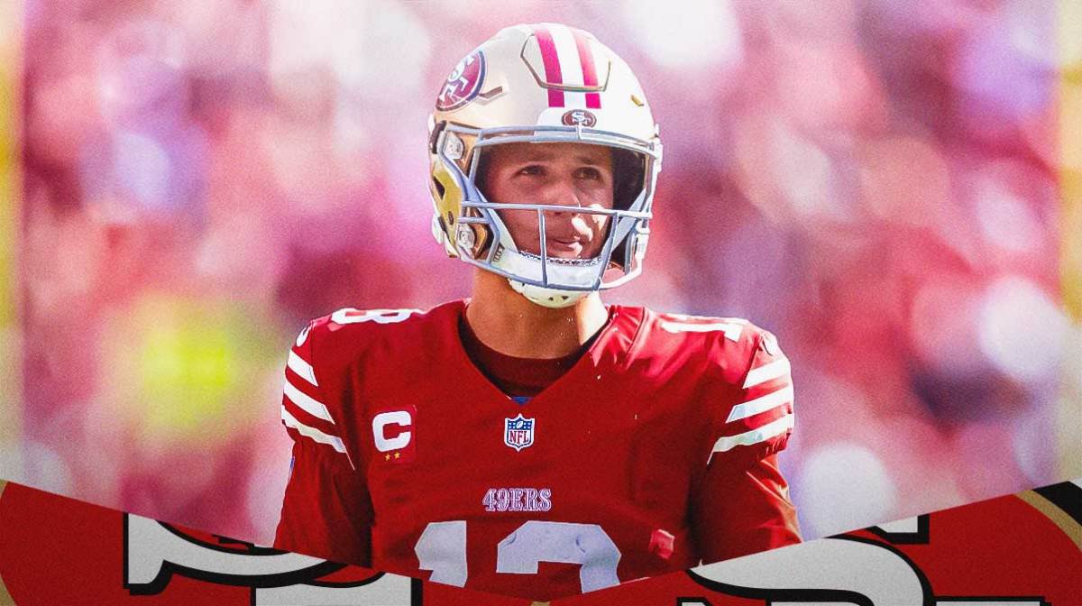 Brock Purdy of the San Francisco 49ers looks on at a game with the 49ers logo underneath.