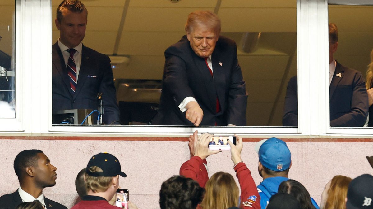President Donald Trump gestures to fans while standing next to U.S. Secretary of War Pete Hegseth (L) after swearing in a new group of U.S. military recruits during halftime of the game between the Detroit Lions and Washington Commanders as part of the NFL Salute to Service initiative at Northwest Stadium.