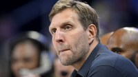 Dirk Nowitzki watches the game between the Dallas Wings and the Seattle Storm during the first half at College Park Center with the Mavericks logo in the background